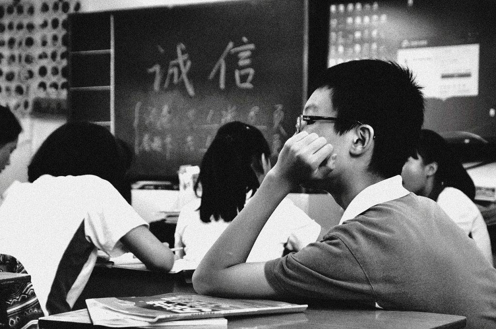 Students in a classroom, one student thoughtfully rests their chin on their hand. Blackboard shows Chinese characters.