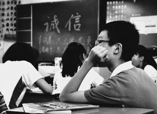 Bina Kuiz Dengan Perisian Mudah Students in a classroom, one student thoughtfully rests their chin on their hand. Blackboard shows Chinese characters.