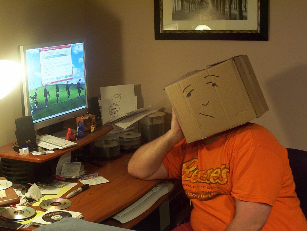 Person with cardboard box on head sits at messy computer desk, looking at screen showing slow computer message. Lajukan Komputer.