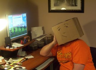Lajukan Komputer Yang Lambat Person with cardboard box on head sits at messy computer desk, looking at screen showing slow computer message. Lajukan Komputer.