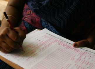Semak Kertas Jawapan OMR dengan Mudah Close-up of hands filling out an OMR answer sheet with a red pencil.